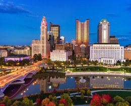 Aerial of Downtown Columbus skyline at dusk during autumn with sunset reflections on the buildings, and with the Scioto River, a bridge, and a park with colorful trees in the foreground.