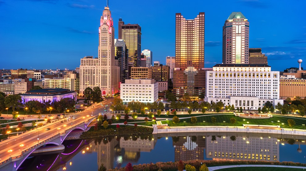 Aerial of Downtown Columbus skyline at dusk during autumn with sunset reflections on the buildings, and with the Scioto River, a bridge, and a park with colorful trees in the foreground.