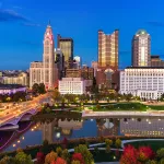 Aerial of Downtown Columbus skyline at dusk during autumn with sunset reflections on the buildings, and with the Scioto River, a bridge, and a park with colorful trees in the foreground.