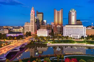 Aerial of Downtown Columbus skyline at dusk during autumn with sunset reflections on the buildings, and with the Scioto River, a bridge, and a park with colorful trees in the foreground.