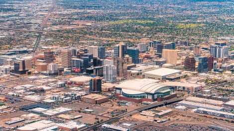 Aerial view of downtown Phoenix, Arizona, with high-rise buildings and a street grid stretching toward hazy mountains.
